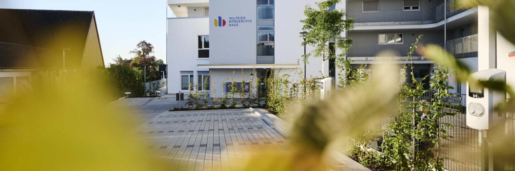 Modernes Mehrfamilienhaus mit weißer Fassade, umgeben von Grün. Am Eingang befindet sich ein Schild mit einem farbenfrohen Logo. Die Szenerie ist an einem klaren Tag aufgenommen, ein gepflasterter Weg führt zur Tür.