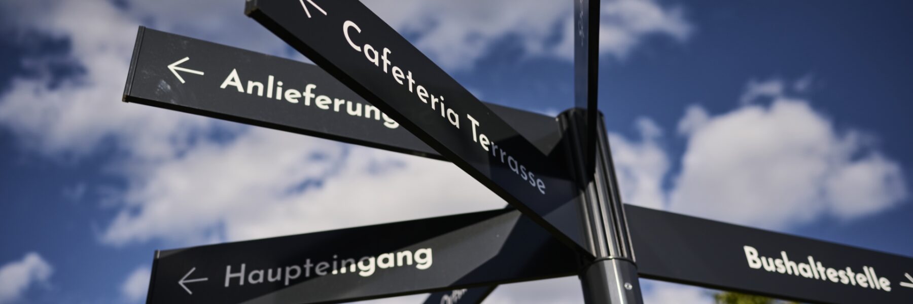 Ein Wegweiser vor einem wolkenverhangenen blauen Himmel mit mehreren schwarzen Pfeilen, die in verschiedene Richtungen zeigen, mit der Aufschrift „Cafeteria Terrasse“, „Anlieferung“, „Haupteingang“ und „Garderobe“.
