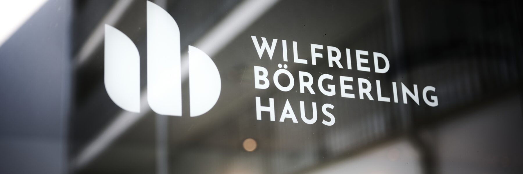 Auf einem Fenster steht der Text „Wilfried Börgerling Haus“ und links davon ein modernes, abstraktes Logo aus drei sich überlappenden Formen. Im Hintergrund ist das verschwommene Innere eines Gebäudes zu sehen.