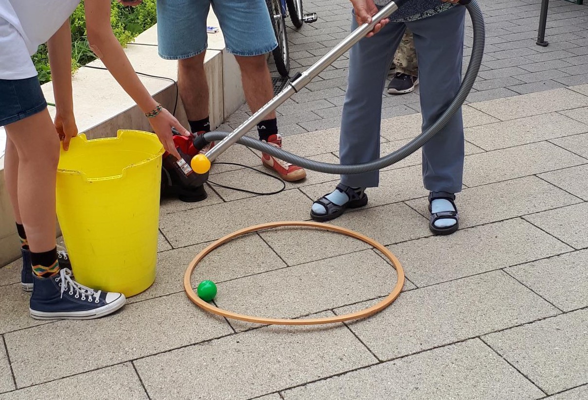 Eine Gruppe von Leuten spielt draußen ein Spiel. Dabei benutzen sie einen Staubsauger, um einen kleinen Ball durch einen Holzreifen auf dem Boden zu schieben. Eine Person hält einen gelben Eimer neben sich.