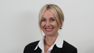 Eine Frau mit blondem Haar, die einen schwarzen Blazer, ein weißes Hemd mit Kragen, eine goldene Halskette und Creolen trägt, lächelt, während sie vor einem schlichten weißen Hintergrund steht.
