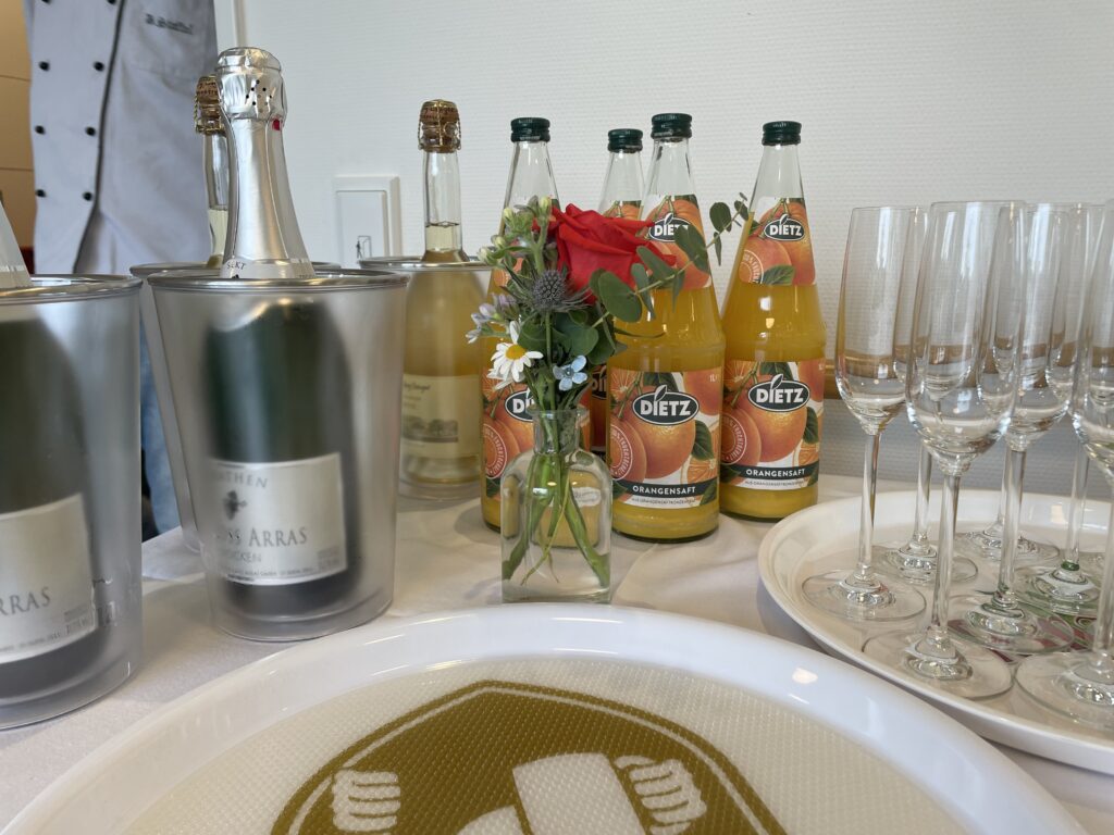 Sekt- und Orangensaftflaschen, eine kleine Blumenvase und leere Sektflöten stehen auf einem weißen Tischtuch, bereit für eine Feier.