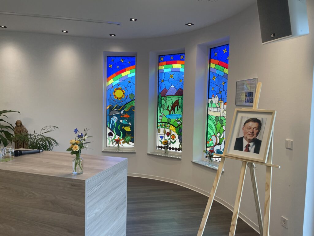 Ein heller Raum mit drei bunten Glasfenstern, einem hellen hölzernen Empfangstisch mit einer Blumenvase und dem gerahmten Foto eines älteren Mannes auf einer Staffelei.