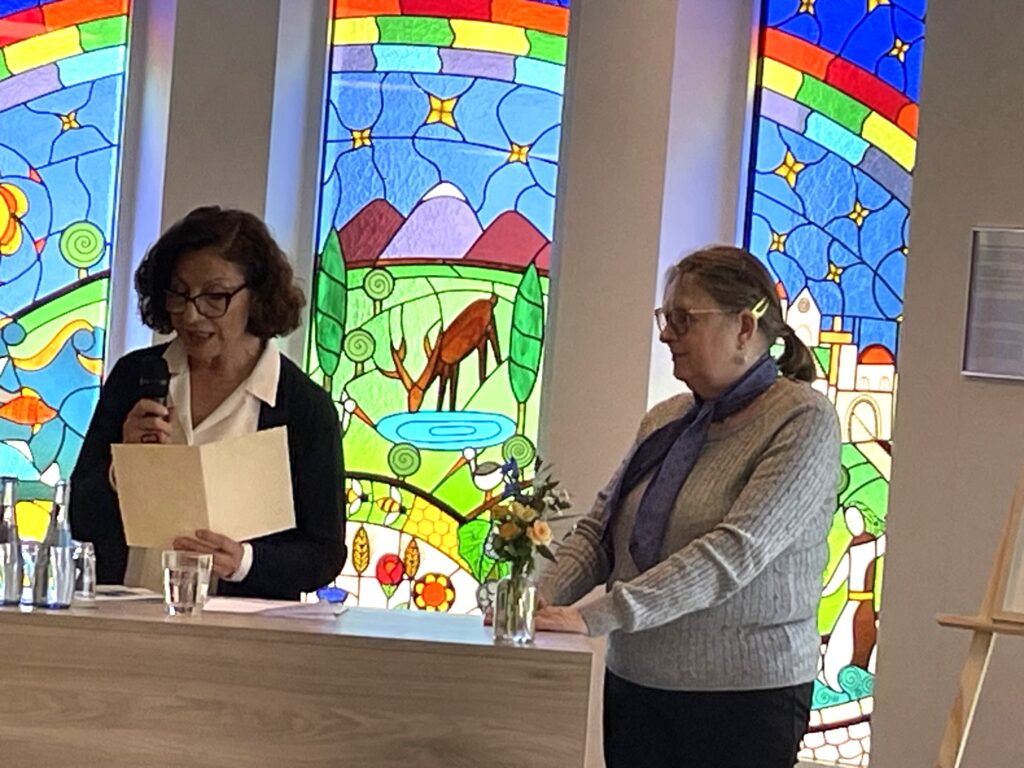 Zwei Frauen stehen an einem Podium; die eine liest von einem Papier in ein Mikrofon, während die andere zuhört. Hinter ihnen sind bunte Glasfenster zu sehen, die ein Reh, eine Landschaft und einen Regenbogen zeigen. Ein kleiner Blumenstrauß steht auf dem Podium.