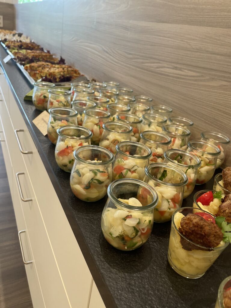 Auf einer Arbeitsplatte stehen reihenweise kleine Gläser, gefüllt mit einem bunten Gemüsesalat, und im Hintergrund ist weiteres Fingerfood zu sehen.