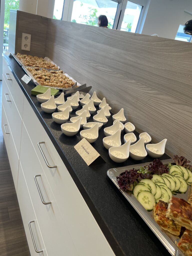 Eine Buffet-Theke mit kleinen weißen Schalen mit cremigem Dip, ein Tablett mit Gurkenscheiben und Grünzeug sowie eine Auswahl an Gebäck in einem modernen Raum mit Tageslicht.