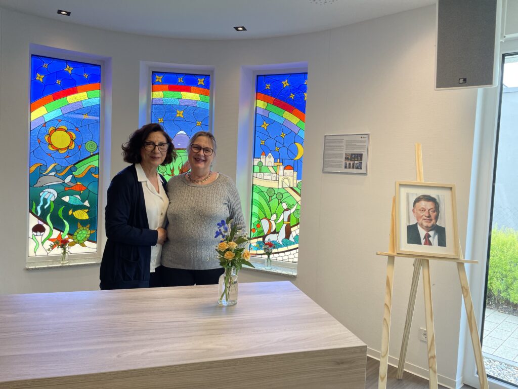 Zwei Frauen stehen lächelnd in einem hell erleuchteten Raum mit bunten Buntglasfenstern, die Regenbögen und Naturszenen zeigen. Ein Tisch mit Blumen und ein gerahmtes Porträt eines Mannes auf einer Staffelei sind ebenfalls zu sehen.
