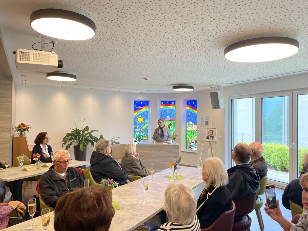 Eine Gruppe älterer Menschen sitzt in einem hellen Raum an Tischen und hört einem Redner auf einem Podium zu. Bunte Buntglasfenster und Blumen schmücken den Raum. Einige der Anwesenden machen Fotos oder halten Getränke in der Hand.