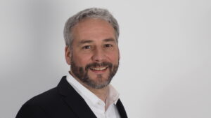 Ein Mann mit kurzen grauen Haaren und einem Bart, der eine schwarze Anzugsjacke und ein weißes Hemd trägt, lächelt in die Kamera vor einem schlichten weißen Hintergrund.